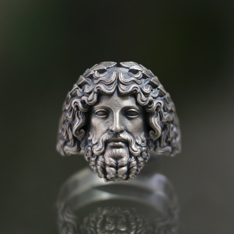 May include: A silver ring with a detailed sculpted face of a man with a beard and a laurel wreath. The ring is a statement piece with a classic design.