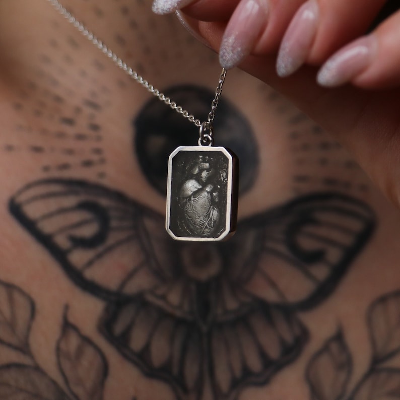 May include: A silver necklace with a rectangular pendant featuring a detailed anatomical heart image.