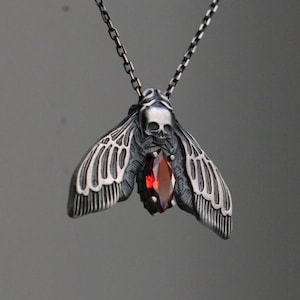 May include: A silver-toned pendant necklace featuring a moth design with a skull at the head and a red gemstone at the center. The moth's wings are detailed with etched lines, and the chain is a linked design.