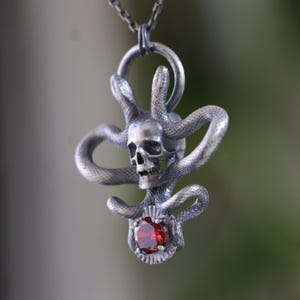 May include: A silver pendant necklace featuring a skull with snakes wrapped around it. A red gemstone is set in the center of the pendant.