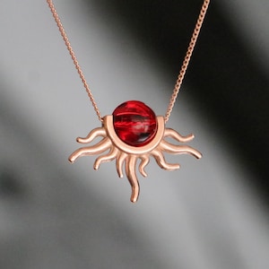 Puede incluir: Un collar de oro rosa con un colgante en forma de sol. El colgante tiene una gema roja redonda central y rayos de sol. La cadena del collar es delicada y de oro rosa.