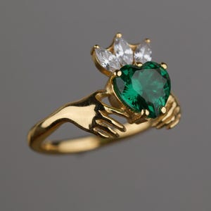 Peut inclure: Une bague dorée avec une pierre précieuse verte en forme de cœur, tenue par deux mains. Au-dessus du cœur se trouve une couronne de pierres précieuses blanches. La bague est posée sur un fond gris.