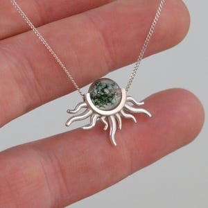K&ouml;nnte beinhalten: Silberkette mit einem sonnenf&ouml;rmigen Anh&auml;nger. Der Anh&auml;nger hat eine klare Glaskugel mit gr&uuml;nen und wei&szlig;en Einschl&uuml;ssen, die in einem silbernen Sonnendesign gefasst ist. Die Halskette ist filigran und silberfarben.