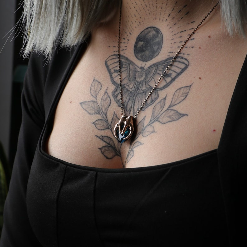 May include: A silver necklace with a blue crystal pendant shaped like a hand holding a heart. The necklace is worn by a person with a butterfly tattoo and a moon tattoo on their chest.