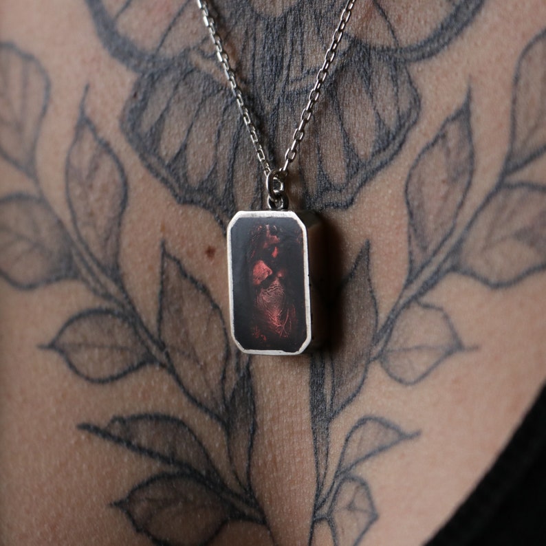 May include: A silver chain necklace with a rectangular pendant featuring a red and black image. The pendant is on a person's chest with a black and gray tattoo.