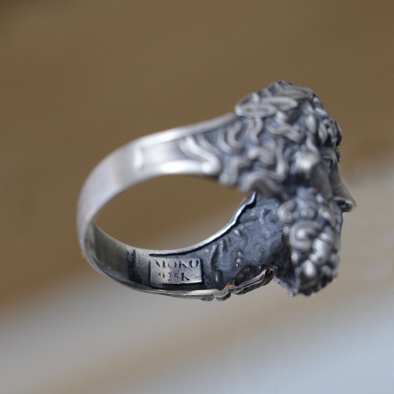 May include: A silver ring with a detailed design of a face with a beard and hair. The ring is marked "MOKO 935K" on the inside.