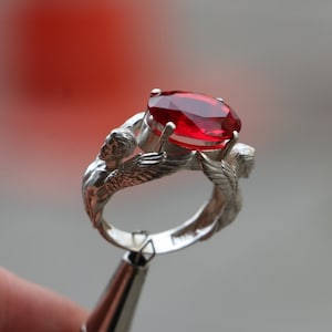 May include: A silver ring with a large, oval-shaped, red gemstone. The ring features two figures with wings on either side of the stone.