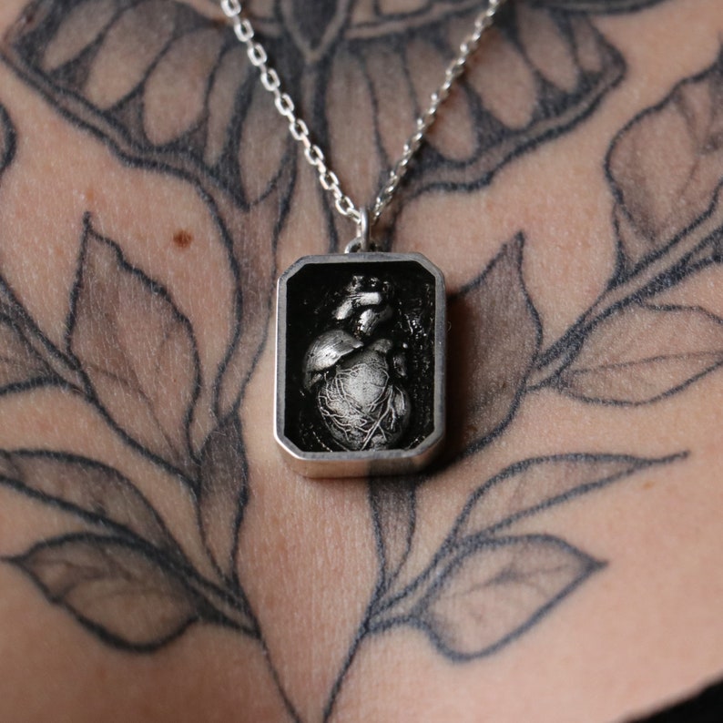 May include: A silver necklace with a rectangular pendant featuring a detailed anatomical heart design.