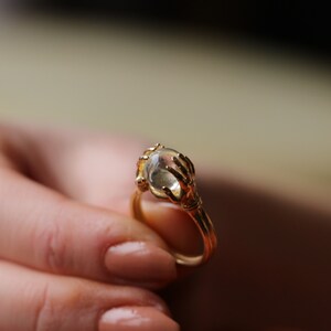 May include: A gold ring featuring two hands clasping a clear glass orb. The ring is a striking piece with an unusual design.