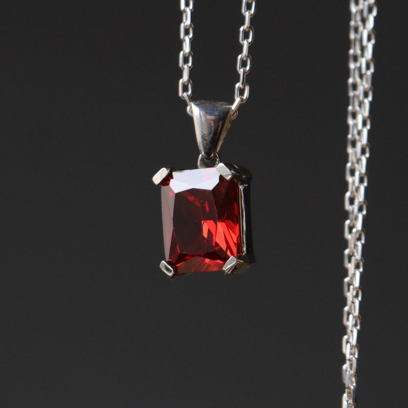 Pu&ograve; includere: Una collana d'argento con un pendente rettangolare di pietra preziosa rosso intenso. Il pendente &egrave; tenuto in posizione da quattro griffe d'argento e pende da una delicata catena d'argento. Lo sfondo &egrave; grigio scuro.