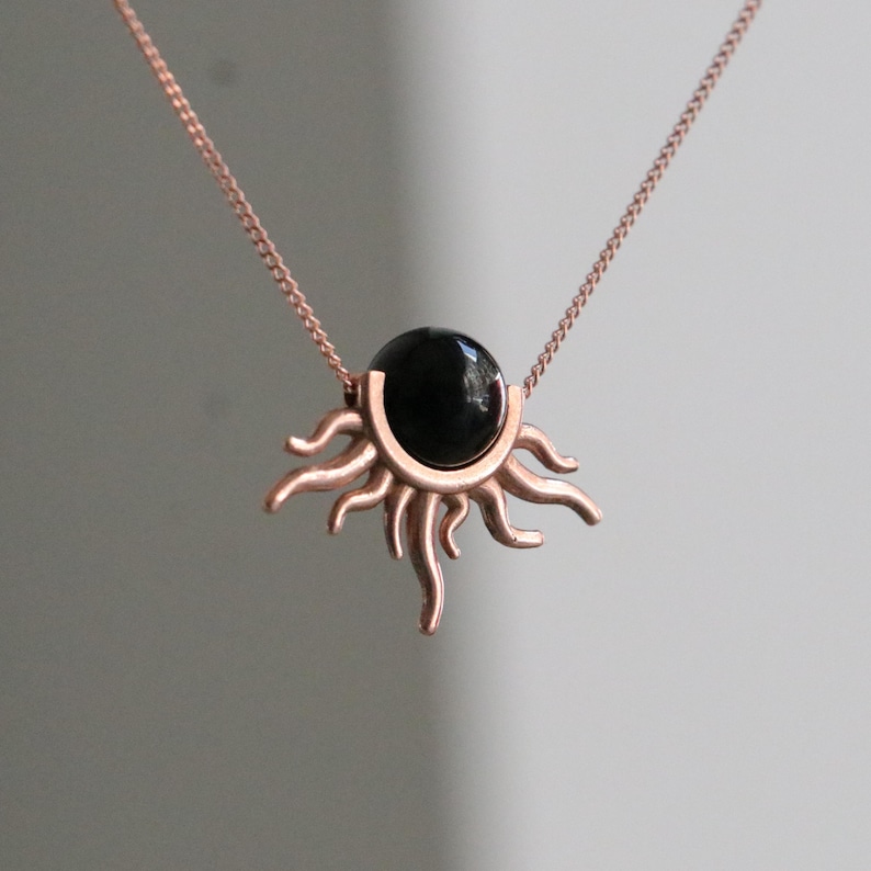 K&ouml;nnte beinhalten: Eine ros&eacute;goldene Halskette mit einem Sonnenanh&auml;nger. Der Anh&auml;nger hat einen zentralen, polierten schwarzen Stein, umgeben von einem Sonnendesign mit stilisierten Strahlen. Die Kette ist zart und erg&auml;nzt das Design des Anh&auml;ngers.