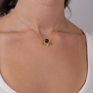 K&ouml;nnte beinhalten: Goldfarbene Halskette mit Anh&auml;nger, der einen dunkelroten Edelstein in der Mitte hat. Der Anh&auml;nger hat ein Sonnenmuster mit geschwungenen Strahlen. Die Halskette wird &uuml;ber einem wei&szlig;en Oberteil getragen.