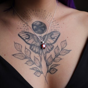 Pu&ograve; includere: Una collana d'argento con un pendente rettangolare di pietra preziosa rosso intenso. Il pendente &egrave; appeso a una catena sottile. La collana &egrave; indossata su uno sfondo di un tatuaggio con una falena e elementi botanici.