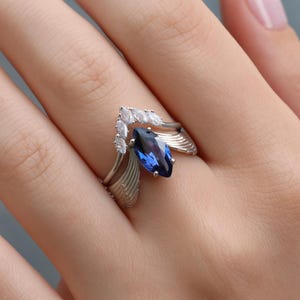 May include: A silver ring with a striking, marquise-cut, deep blue gemstone. The ring's design includes a cluster of small, clear stones and wing-like details. The ring is displayed on a person's finger.