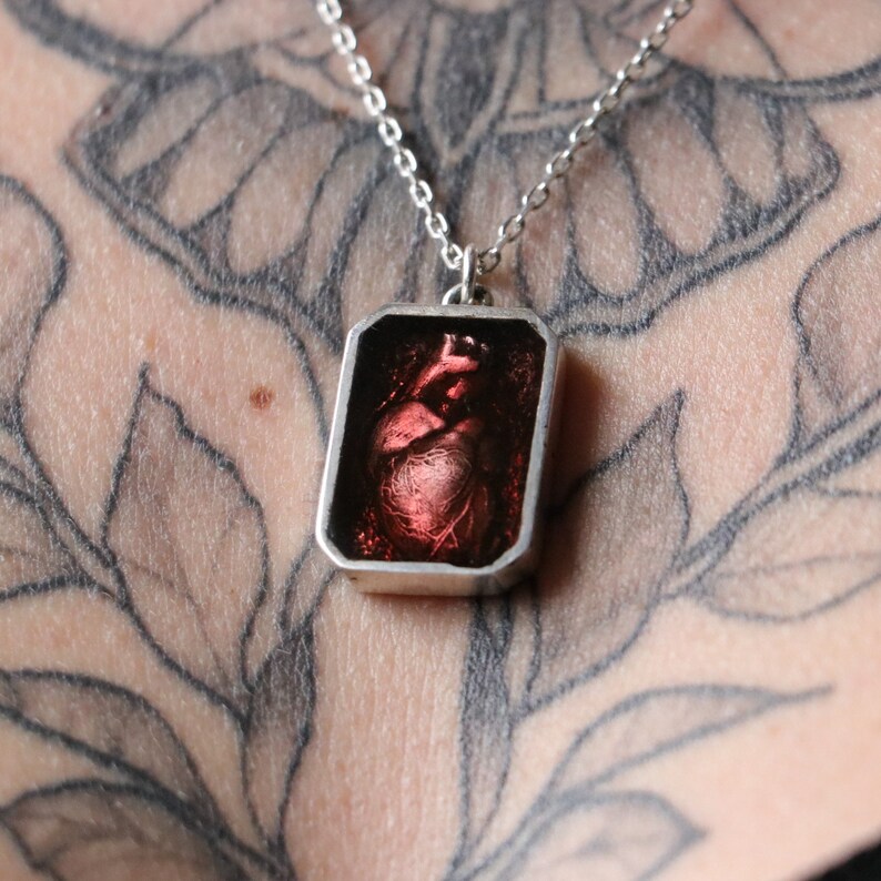 May include: A silver chain necklace with a rectangular pendant featuring a red anatomical heart.