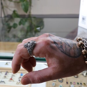 May include: A silver ring with a detailed design of a face with a beard. The ring is worn on the index finger of a hand with a tattoo.