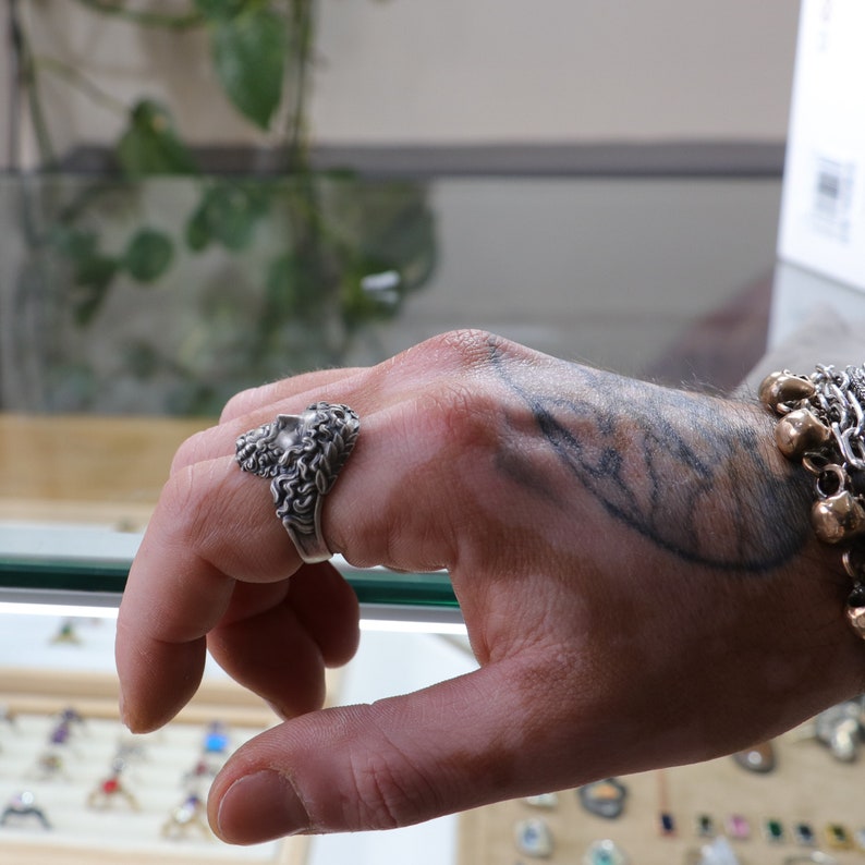 May include: A silver ring with a detailed design of a face with a beard. The ring is worn on the index finger of a hand with a tattoo.