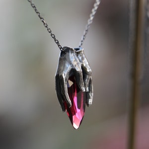 May include: A silver necklace with a pendant featuring two hands holding a large, faceted, dark red gemstone.