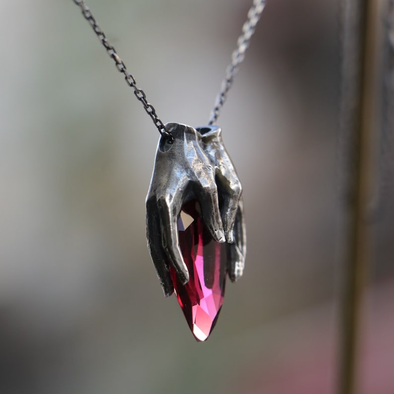 May include: A silver necklace with a pendant featuring two hands holding a large, faceted, dark red gemstone.