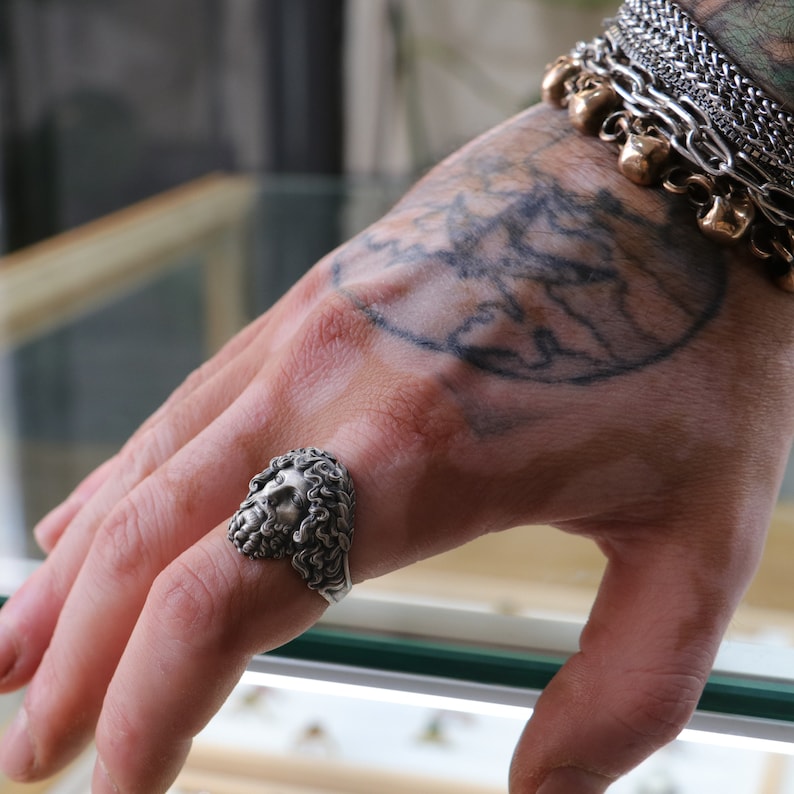 May include: A silver ring with a detailed sculpted face design. The ring is worn on the index finger of a hand with a tattoo of a world map.
