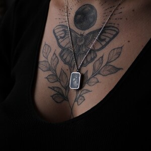 May include: A silver necklace with a rectangular pendant featuring a black and white image. The necklace is worn by a person with a butterfly and moon tattoo on their chest.