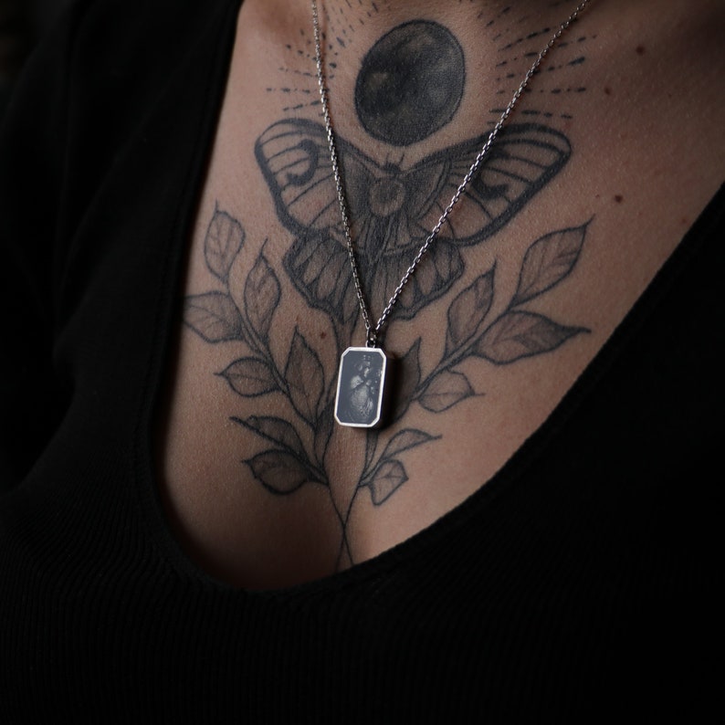 May include: A silver necklace with a rectangular pendant featuring a black and white image. The necklace is worn by a person with a butterfly and moon tattoo on their chest.