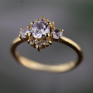 May include: A gold ring featuring a large, round, clear gemstone at the center, surrounded by smaller clear stones. The ring has a delicate, detailed setting and a smooth, polished band.