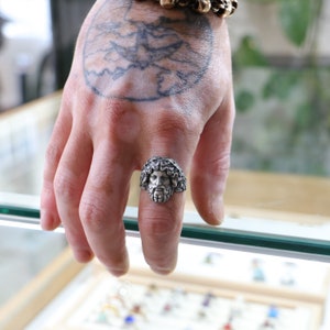 May include: A silver ring with a detailed sculpted face of a man with curly hair. The ring is worn on the index finger of a hand with a tattoo.