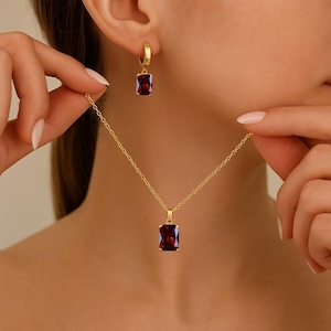Peut inclure: Un ensemble de bijoux de couleur or comprenant un collier et des boucles d'oreilles. Le collier est orné d'un pendentif rectangulaire en pierre précieuse rouge foncé. Les boucles d'oreilles sont de style créole, chacune avec une pierre précieuse assortie.