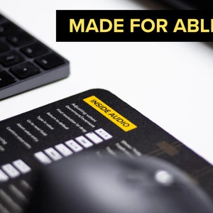 May include: A black mouse pad with a yellow label that says "INSIDE AUDIO" and the text "MADE FOR ABLETON" in black letters. The mouse pad has a grid pattern and a keyboard and mouse are on the surface.