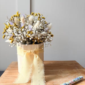 May include: A bouquet of dried flowers in yellow, white, and light purple hues arranged in a light-coloured wooden container. A pale yellow ribbon with the text "thank you" printed in small letters is tied around the container. A floral patterned pen.