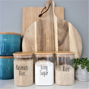 May include: Three glass jars with bamboo lids, labeled "Basmati Rice", "Icing Sugar", and "Rice". The jars are filled with different types of rice and sugar. The jars are sitting on a counter in front of a wooden cutting board.
