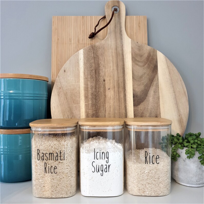 May include: Three glass jars with bamboo lids, labeled "Basmati Rice", "Icing Sugar", and "Rice". The jars are filled with different types of rice and sugar. The jars are sitting on a counter in front of a wooden cutting board.
