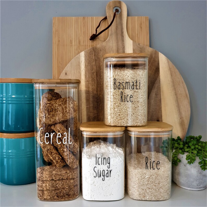 May include: Five glass jars with bamboo lids, labeled with handwritten labels. The labels read "Cereal", "Basmati Rice", "Rice", "Icing Sugar", and "Rice". The jars are filled with various pantry staples, including cereal, rice, and powdered sugar.