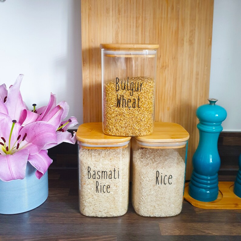 May include: Three glass jars with bamboo lids filled with different types of rice. The jars are labeled "Bulgur Wheat", "Basmati Rice", and "Rice".