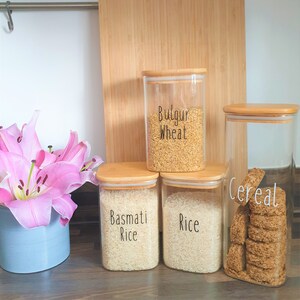 May include: Four glass jars with bamboo lids, filled with various grains and cereal. The jars are labeled "Bulgur Wheat", "Basmati Rice", "Rice", and "Cereal".