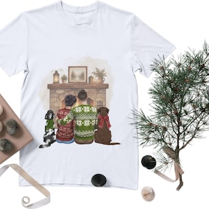 May include: A white t-shirt featuring a graphic illustration of two people sitting in front of a stone fireplace. The people wear patterned sweaters, and two dogs sit beside them. The fireplace mantel holds a framed picture, a lantern, and plants.