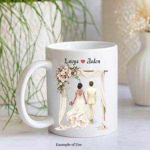 May include: A white ceramic mug featuring a printed illustration of a couple standing under a floral wedding arch. The text Latoya and Jaden is printed above the couple, separated by a red heart. The couple is shown from behind, holding hands.