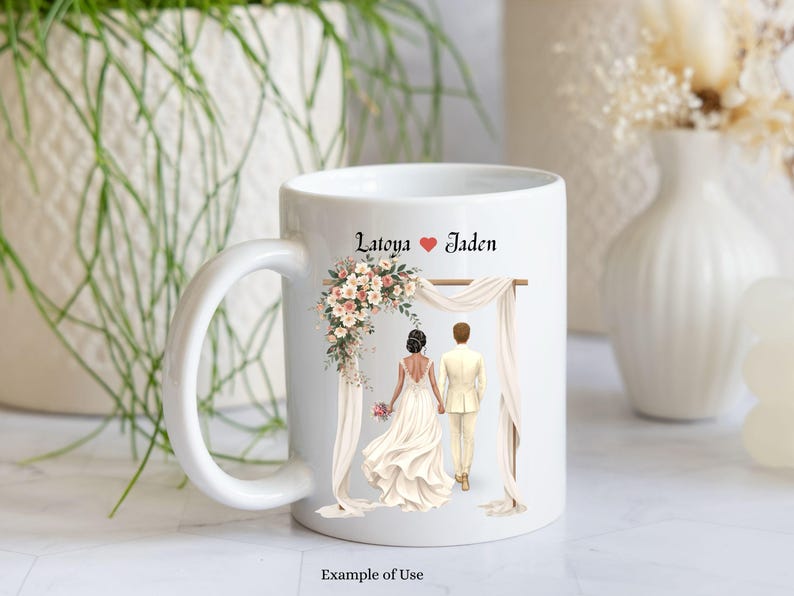 May include: A white ceramic mug featuring a printed illustration of a couple standing under a floral wedding arch. The text Latoya and Jaden is printed above the couple, separated by a red heart. The couple is shown from behind, holding hands.