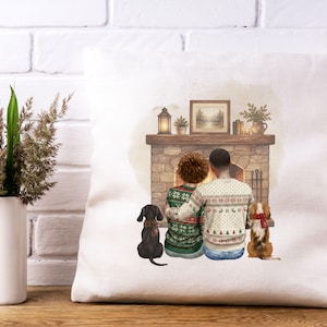 May include: A square white throw pillow featuring a printed illustration of two people sitting together in front of a stone fireplace. They wear patterned holiday sweaters, and two dogs sit beside them, one black and one brown, both wearing scarves.