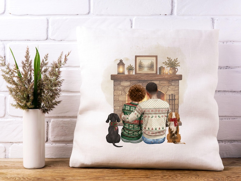 May include: A square white throw pillow featuring a printed illustration of two people sitting together in front of a stone fireplace. They wear patterned holiday sweaters, and two dogs sit beside them, one black and one brown, both wearing scarves.
