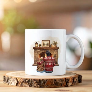 May include: A white ceramic mug featuring a printed illustration of two people sitting in front of a stone fireplace. The individuals are wearing patterned red and beige sweaters. The fireplace mantel holds a framed picture, a lantern, and a plant.
