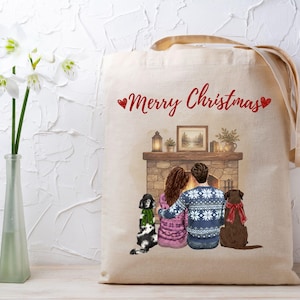 May include: A beige canvas tote bag featuring a printed illustration of two people and two dogs sitting before a fireplace. The text Merry Christmas is printed in red above the scene. The people wear patterned sweaters and the dogs wear scarves.