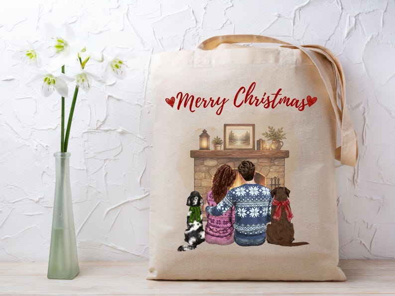May include: A beige canvas tote bag featuring a printed illustration of two people and two dogs sitting before a fireplace. The text Merry Christmas is printed in red above the scene. The people wear patterned sweaters and the dogs wear scarves.