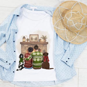 May include: A white t-shirt featuring a graphic illustration of two people and two dogs sitting before a stone fireplace. The people wear patterned sweaters, and the dogs wear festive scarves. A light blue button-down shirt and a straw hat are included.
