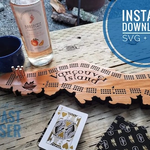 Op de afbeelding: Een houten spelbord in de vorm van Vancouver Island met de tekst "Vancouver Island" erop gedrukt. Het bord heeft een raster van gaten om een spel te spelen. Het bord ligt op een houten tafel met een glas perzikwijn, een blauwe mok en een kaartspel.