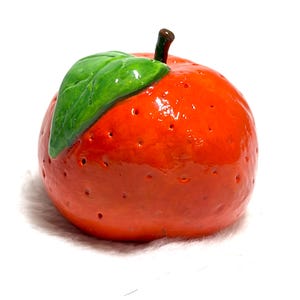 May include: A glossy, bright orange citrus fruit figurine with a textured surface featuring small indentations. A single, smooth, vibrant green leaf is attached to the top, next to a small, dark brown stem. The object has a rounded, spherical shape.