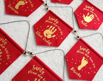 May include: A collection of red felt pennant banners with gold lettering that reads Santa please stop here for, featuring gold handprints or footprints and personalised names including Tiennan, Paudi, Arlo, Elsie Violet, Brooke, and Sidney Job.