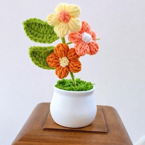 Può includere: Un'arrangiamento floreale all'uncinetto fatto a mano in un vaso di ceramica bianco. I fiori sono nei toni del giallo, arancione e pesca con foglie verdi. Il vaso è posto su una superficie di legno, creando un'affascinante decorazione.