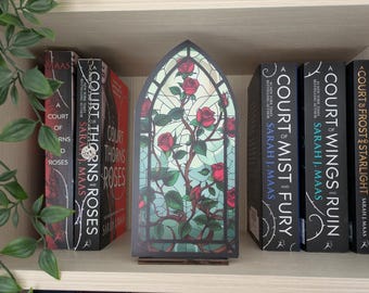 May include: A bookshelf with several books and a decorative stained glass panel. The books are titled "Court of Thorns and Roses", "Court of Mist and Fury", "Court of Wings and Ruin", and "Court and Frost and Starlight". The panel features red roses.